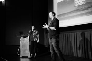 Black and white photo of two people talking to audience on stage