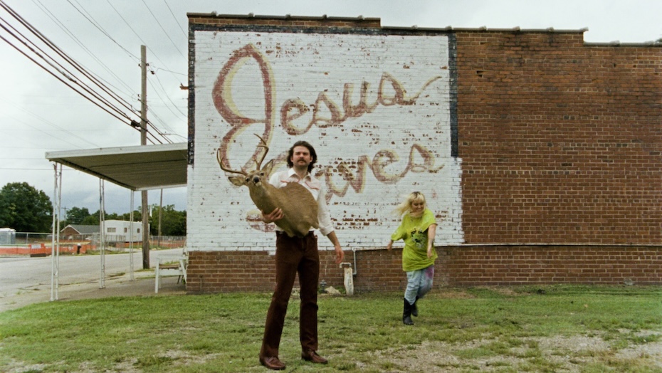 Image of person holding taxidermy deer head outside while another person dances.