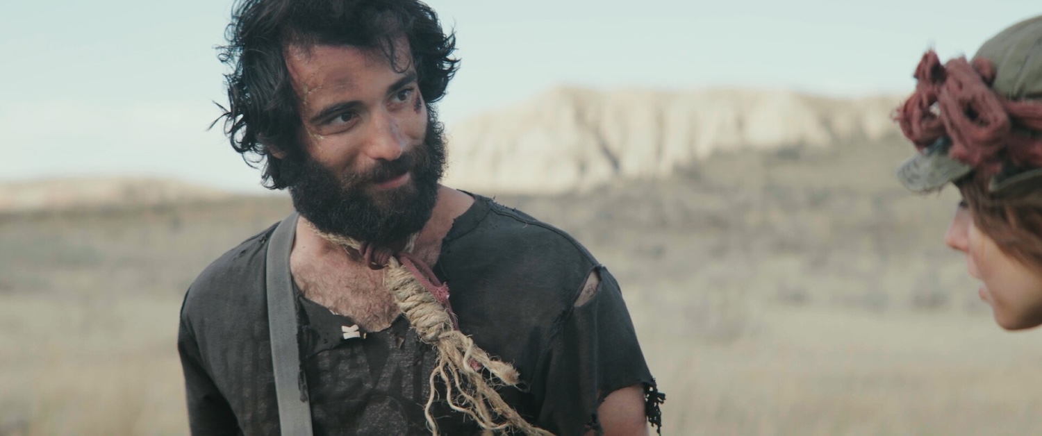 Image of person with noose around neck looks at another person in dry landscape.